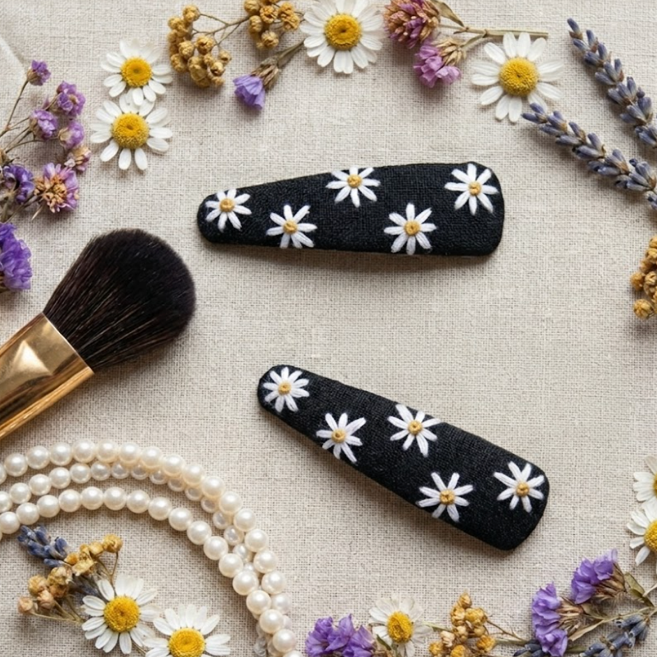 Black hair clips with white daisy patterns, a makeup brush, and dried flowers on a textured surface.