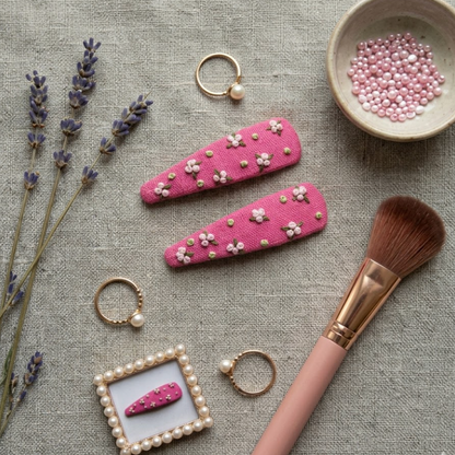 Pink hair clips with floral details on a textured surface with lavender, a brush, and jewelry.